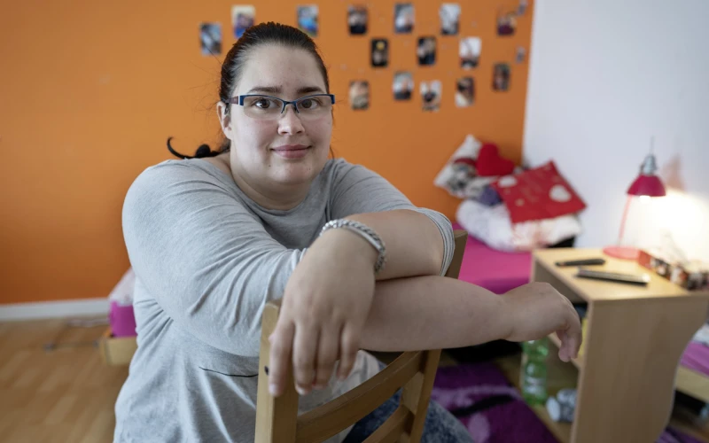 Eine Frau mit Brille sitzt entspannt verkehrt herum auf einem Stuhl in einem Raum mit einer orangefarbenen Wand. Im Hintergrund sind ein Bett, ein kleiner Schrank und eine Collage aus Fotos zu sehen. Sie tr&auml;gt ein graues Oberteil und lehnt sich bequem auf die Stuhllehne.