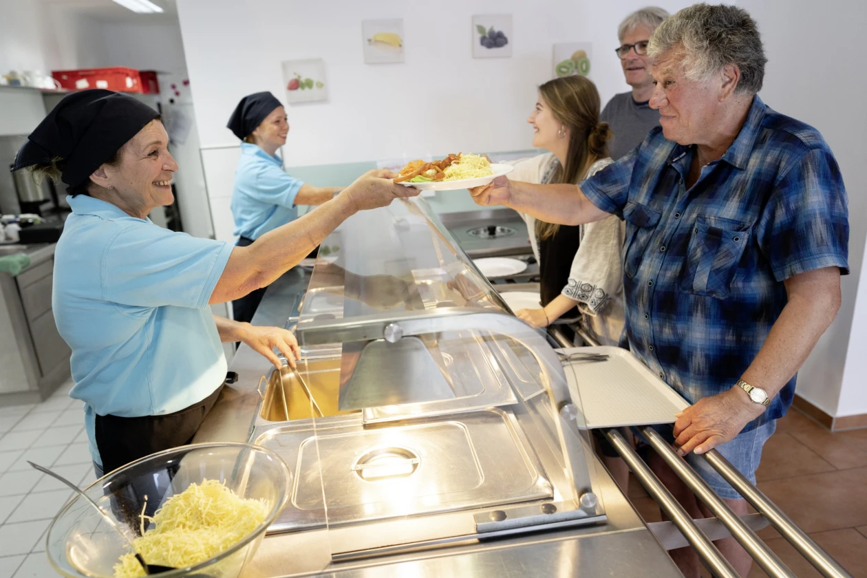 Zwei Frauen in hellblauen T-Shirts geben Essen an G&auml;ste an einer Ausgabetheke aus. Eine Frau reicht einem Mann in kariertem Hemd eine Portion Essen, w&auml;hrend im Hintergrund zwei weitere Personen warten.