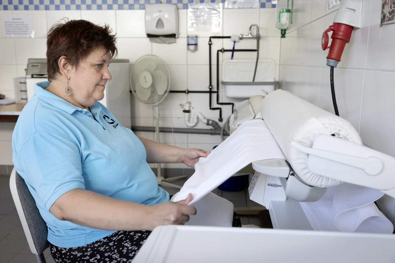 Frau in hellblauen T-Shirt mit Erlacher H&ouml;he Logo sitzt an einer B&uuml;gelmaschine in einer W&auml;scherei und faltet wei&szlig;e W&auml;sche. Ein Ventilator und industrielle Rohrleitungen sind im Hintergrund zu sehen.