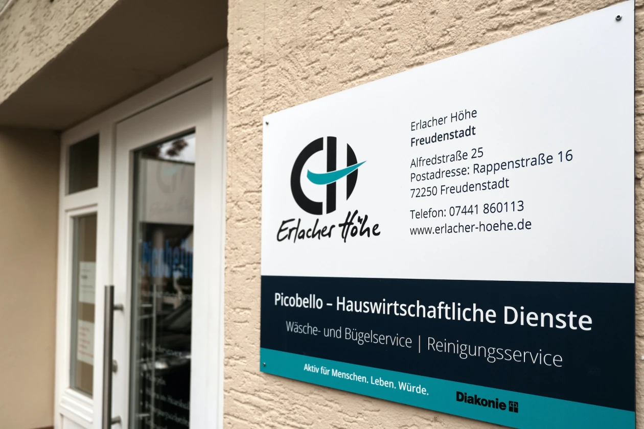 Schild der Erlacher H&ouml;he mit Logo, Kontaktinformationen und Hinweis auf Picobello-Hauswirtschaftliche Dienste, angebracht an einer beigen Hauswand rechts neben einer Eingangst&uuml;r.