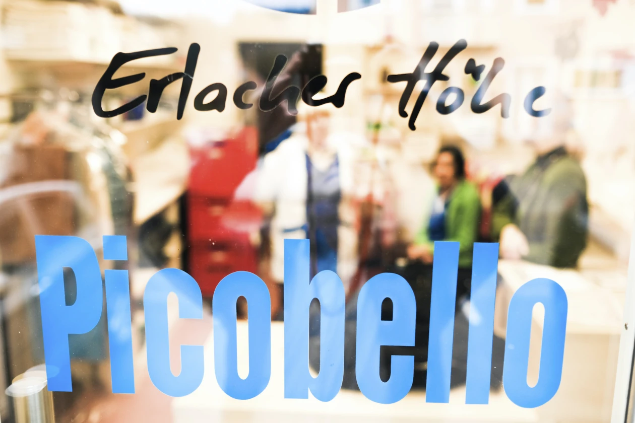 Glast&uuml;re mit dem blauen Schriftzug "Picobello" und dem Text "Erlacher H&ouml;he" in schwarzer Schrift. Beim Durchblicken sind verschwommen Menschen zu sehen.