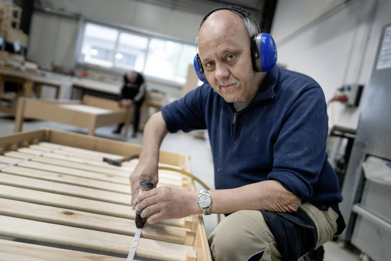 Ein Mann mit blauem Geh&ouml;rschutz l&auml;chelt in die Kamera. Er arbeitet in einer Werkstatt an einem Bettgestell. Im Hintergrund ist eine weitere Person zu sehen, die ebenfalls an einem Bettgestell arbeitet.