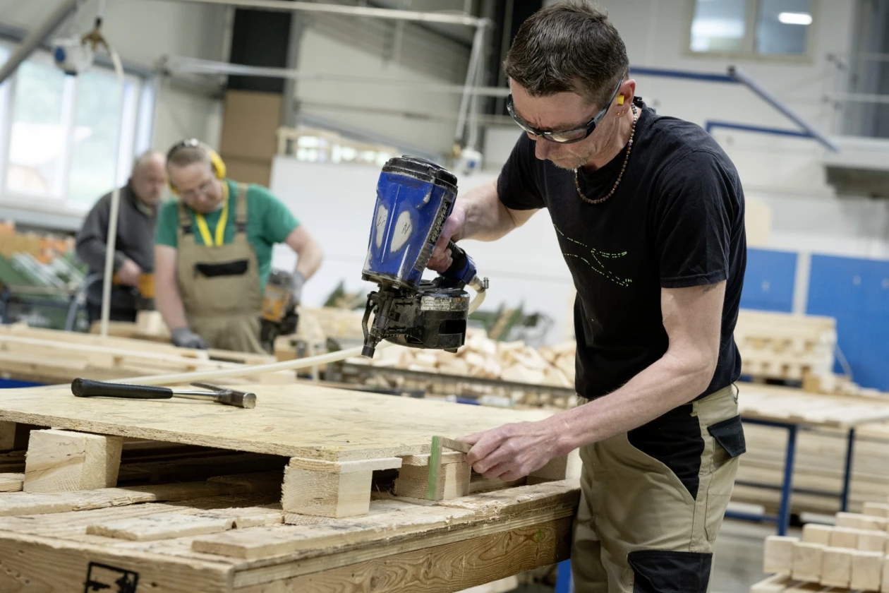 M&auml;nner in einer Werkstatt arbeiten an Holzpaletten. Ein Mann in schwarzem T-Shirt und Brille verwendet einen blauen Druckluftnagler, w&auml;hrend ein anderer Mann im Hintergrund t&auml;tig ist.