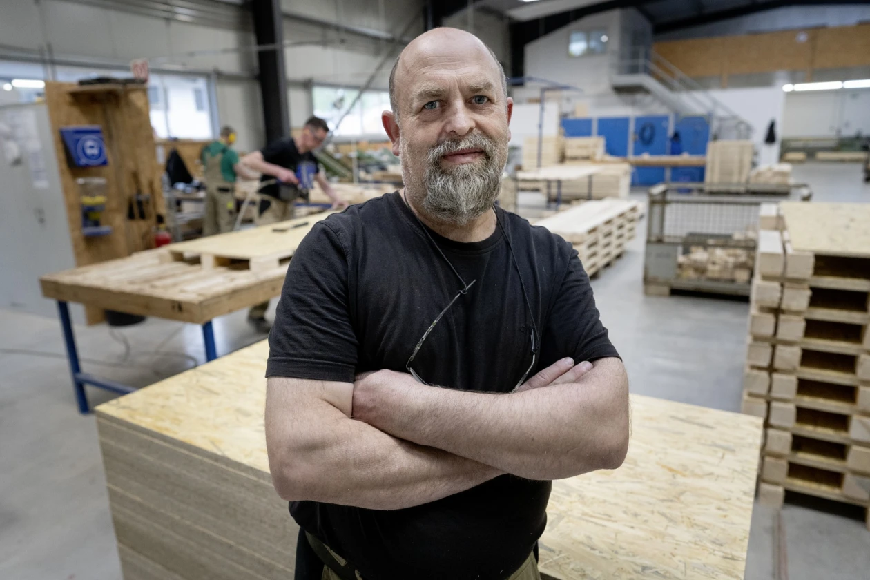 Ein Mann mit grauem Bart und schwarzem T-Shirt steht verschr&auml;nkten Armen in einer Werkstatt mit Holzpaletten und Arbeitstischen im Hintergrund.