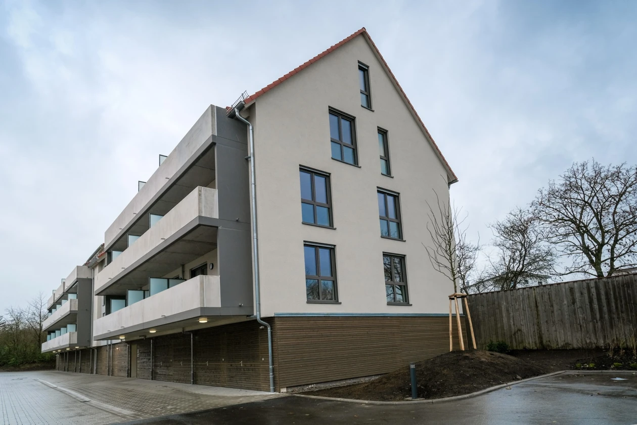 Geb&auml;ude mit drei Etagen in hellen Farben, bestehend aus einem spitzem Dach und einem Seitenfl&uuml;gel mit Balkonen. Das Geb&auml;ude hat eine helle Fassade, einen dunklen Sockel und gro&szlig;e rechteckige Fenster, umgeben von einem gepflasterten Parkplatzbereich und einem Zaun.