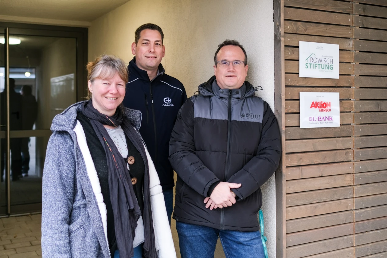 Eine Frau und zwei M&auml;nner stehen im Eingangsbereich und l&auml;cheln in die Kamera.