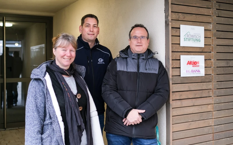 Eine Frau und zwei M&auml;nner stehen im Eingangsbereich und l&auml;cheln in die Kamera.
