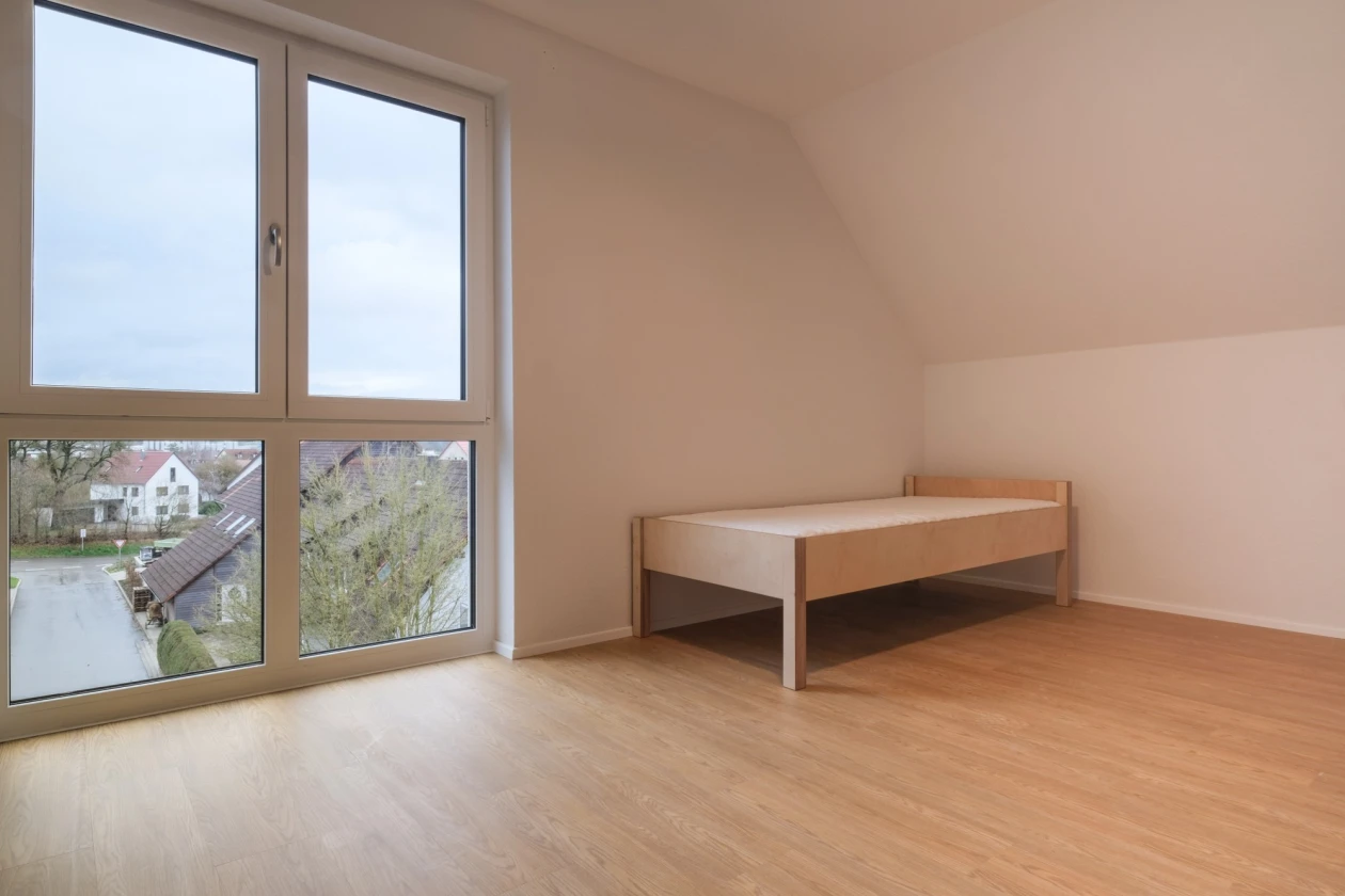 Leeres Zimmer mit Holzboden und einem Einzelbett. Gro&szlig;e Fenster bieten Ausblick auf H&auml;user und Landschaft.