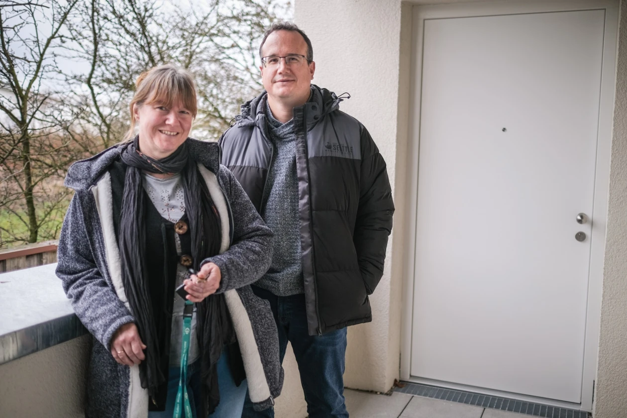 Zwei Personen stehen nebeneinander auf einem Balkon. Beide tragen warme Winterkleidung wie Jacken und Schals. Sie l&auml;cheln freundlich in die Kamera und stehen vor einer wei&szlig;en T&uuml;r mit kahlen B&auml;umen im Hintergrund.