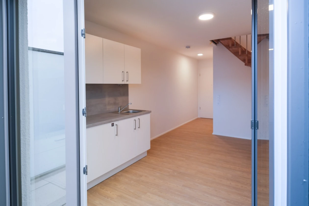 Ein Raum mit Holzboden und einer wei&szlig;-grauen K&uuml;chenzeile. Die Glast&uuml;r steht offen, und im Hintergrund ist eine Treppe sichtbar. Die W&auml;nde sind glatt und wei&szlig;.