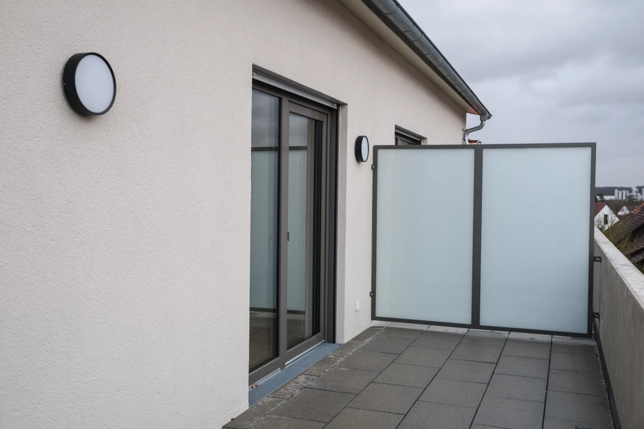 Ein Balkon mit grau gefliester Terrasse, gro&szlig;er bodentiefer Glasfenstert&uuml;r und einem mattgrauen Sichtschutz-Paravent. Zwei schwarze runde Wandleuchten sind an der hellen Hauswand montiert, im Hintergrund sind weitere H&auml;user zu sehen.