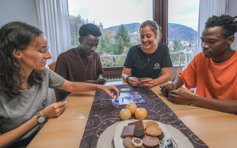 Zwei Frauen und zwei M&auml;nner sitzen an einem Tisch und spielen Mensch &auml;rgere dich nicht, w&auml;hrend im Hintergrund durch ein Fenster Berge und B&auml;ume zu sehen sind. Auf dem Tisch liegt Obst und Geb&auml;ck..
