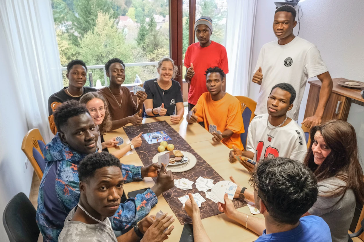 Eine Gruppe von zw&ouml;lf Personen mit unterschiedlichen Hautfarben sitzt um einen Tisch, auf dem Spielkarten, ein Spielbrett f&uuml;r Mensch &auml;rgere dich nicht, Obst und Geb&auml;ck verteilt sind. Der Raum ist hell und hat Fenster, durch die man den Blick ins Gr&uuml;ne genie&szlig;en kann.