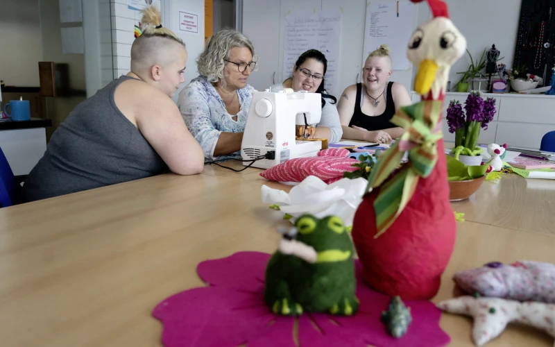 Vier Frauen sitzen an einem Tisch und arbeiten mit einer N&auml;hmaschine, w&auml;hrend im Vordergrund eine rote H&uuml;hnerfigur aus Filz und andere Dekorationsgegenst&auml;nde platziert sind.