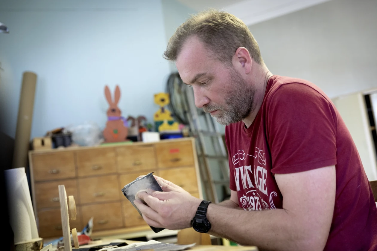 Ein b&auml;rtiger Mann in einem roten T-Shirt konzentriert sich bei der Arbeit in einer Werkstatt und h&auml;lt einen Gegenstand in den H&auml;nden, w&auml;hrend Bastelutensilien und ein Holzschrank im Hintergrund zu sehen sind.