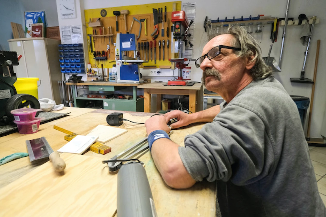 Ein Mann mit Brille arbeitet an einem Tisch in einem Raum mit vielen Werkzeugen und Arbeitsmaterialien.