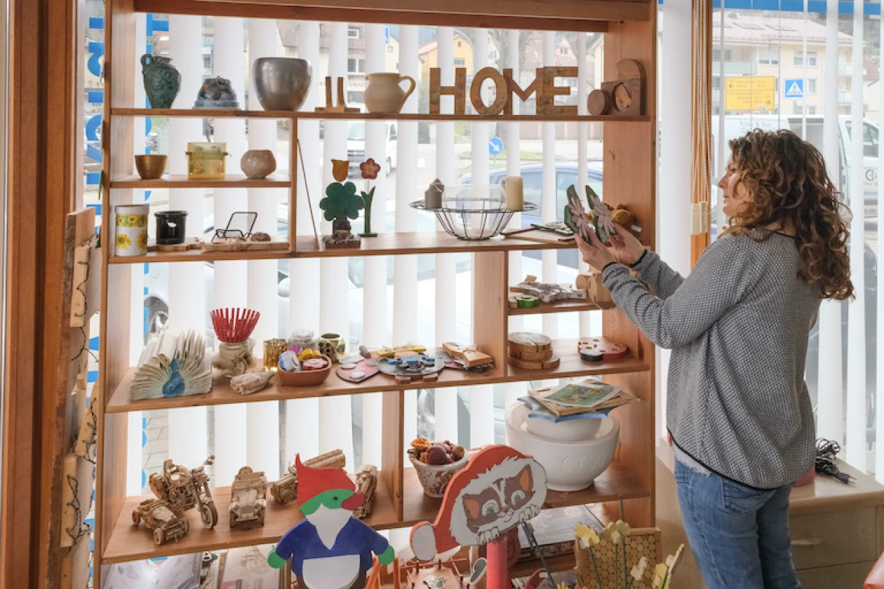 Eine Frau mit lockigem Haar steht vor einem Holzregal mit verschiedenen Keramikgef&auml;&szlig;en, T&ouml;pfen und Dekorationsgegenst&auml;nden. Die Frau h&auml;lt einen Gegenstand in den H&auml;nden und betrachtet ihn pr&uuml;fend.