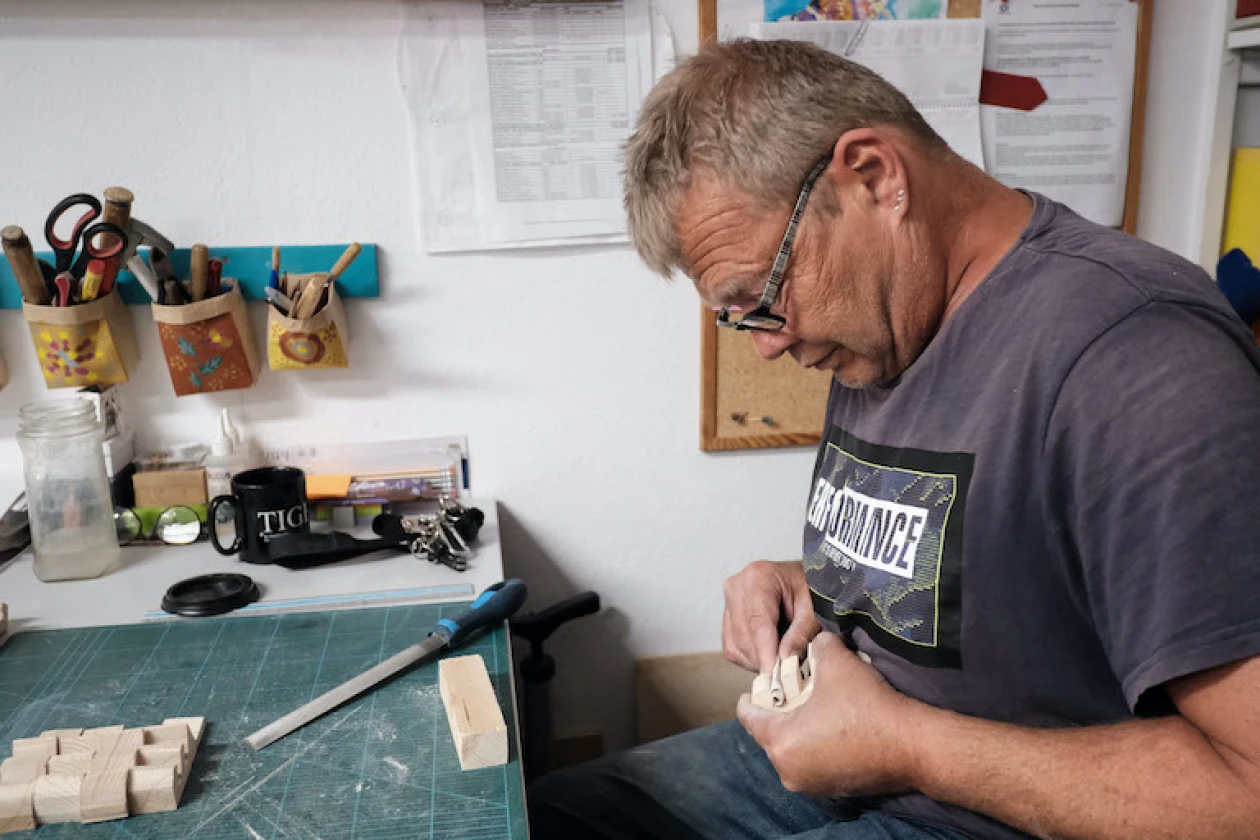 Ein Mann mit Brille arbeitet konzentriert an einem Handwerkstisch und h&auml;lt ein Holzst&uuml;ck in den H&auml;nden. Auf dem Tisch sind weitere Werkzeuge und Materialien zu sehen. Im Hintergrund sieht man eine Pinnwand und Dokumente an der Wand h&auml;ngen.