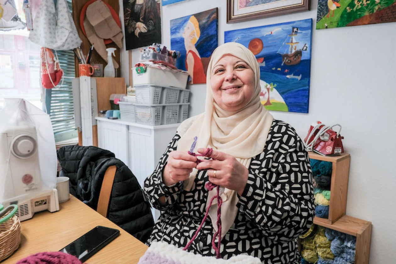 Eine l&auml;chelnde Frau mit Hijab sitzt an einem Tisch, auf dem eine N&auml;hmaschine steht. Sie strickt konzentriert mit Stricknadeln und Wolle. Im Hintergrund sind Kisten mit Bastelmaterialien zu sehen.