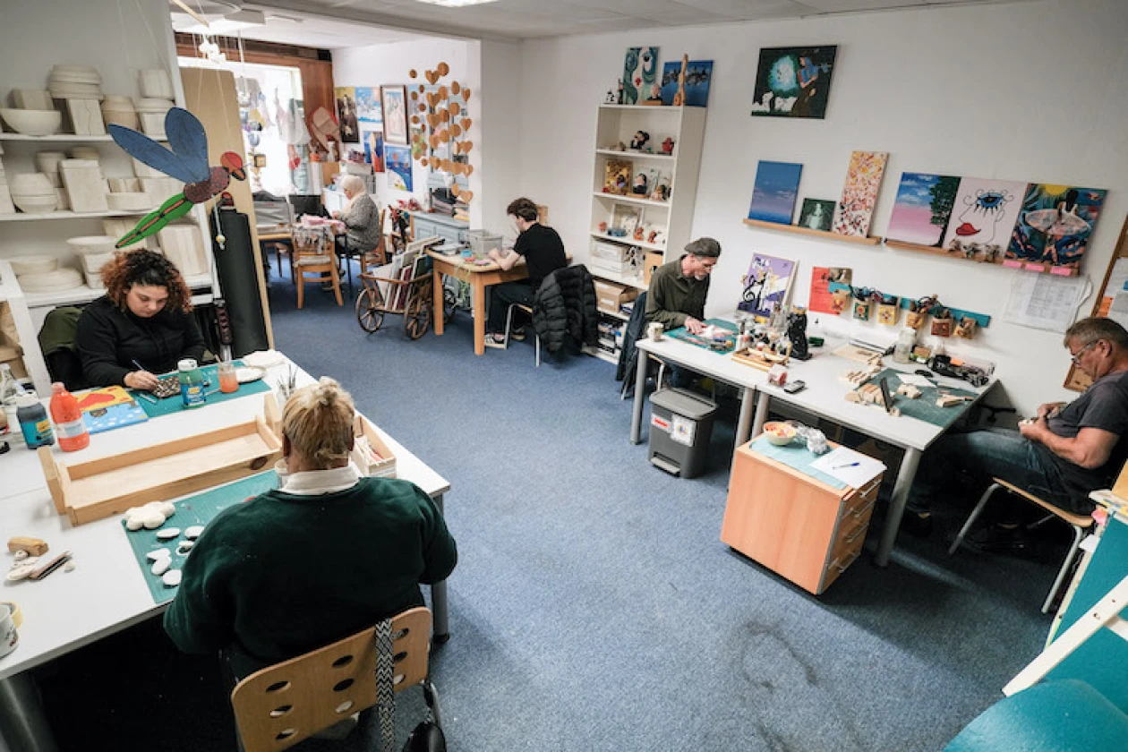 Ein Raum mit mehreren Arbeitspl&auml;tzen, ausgestattet mit Tischen, Regalen und Kunstwerken an den W&auml;nden. Verschiedene Personen arbeiten konzentriert an kreativen Projekten in einem hellen Raum.