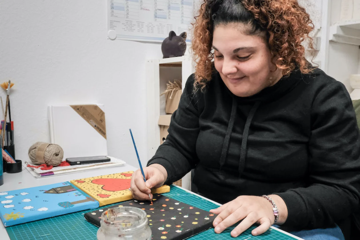 Eine junge Frau mit lockigem Haar sitzt an einem Tisch und tupft mit einem Pinsel Farbe auf eine kleine Leinwand. Sie tr&auml;gt einen schwarzen Pullover und arbeitet auf einer blauen Unterlage. Auf dem Tisch sind weitere fertige Bilder zu sehen.