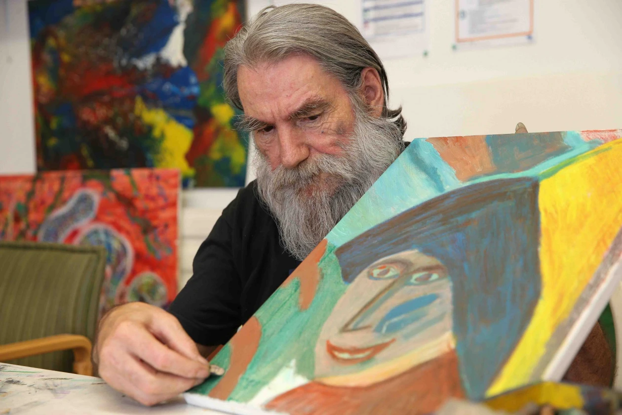 Ein Mann mit einem langen grauen Bart malt an einem abstrakten Gem&auml;lde mit pastellfarbenen T&ouml;nen. Er tr&auml;gt ein schwarzes T-Shirt und im Hintergrund sind weitere Gem&auml;lde zu sehen.