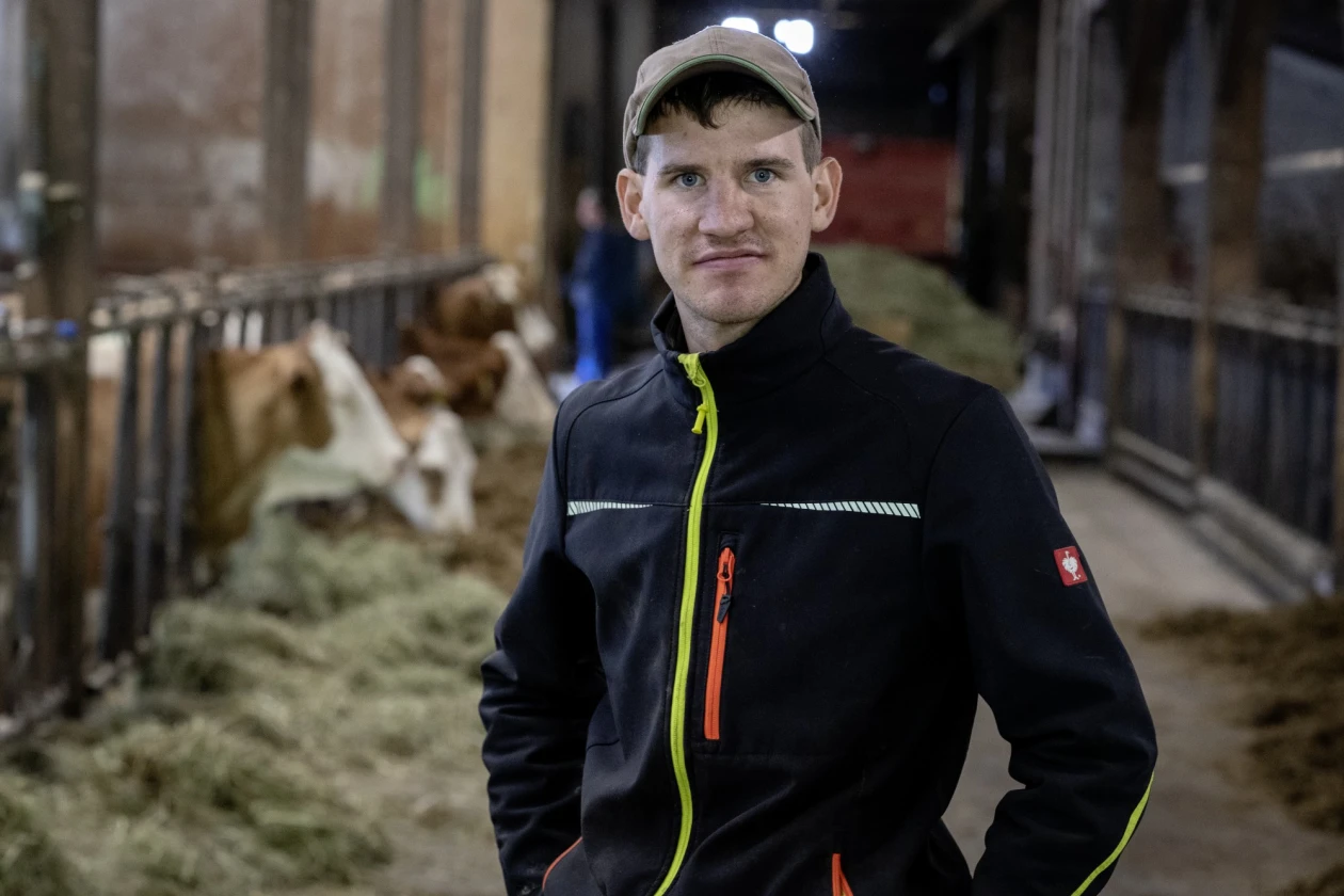 Ein junger Mann in schwarzer Arbeitsjacke mit auff&auml;lligen Rei&szlig;verschl&uuml;ssen steht in einem Kuhstall mit Heu und Rindern im Hintergrund. Er tr&auml;gt eine Kappe und blickt geradeaus in die Kamera.
