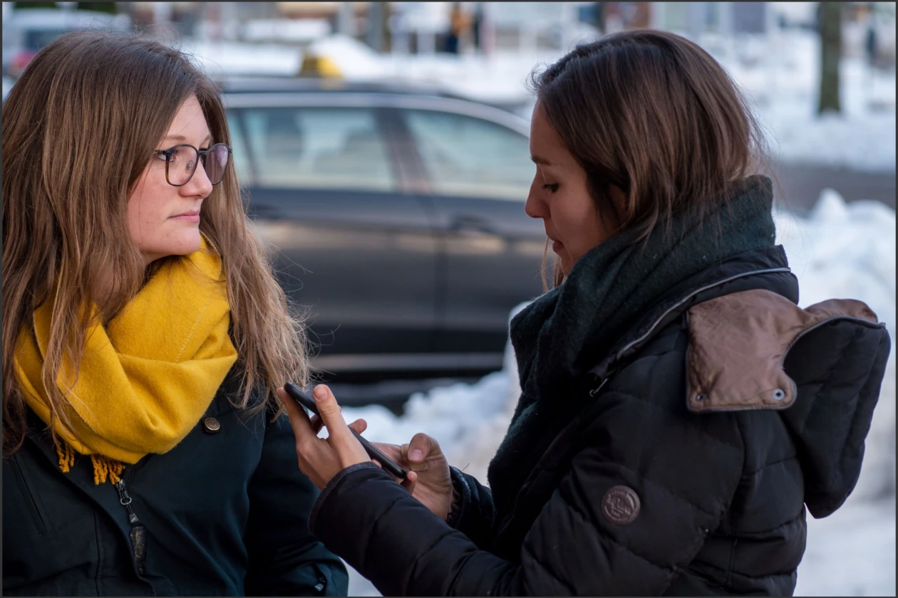 Eine junge Frau tr&auml;gt einen gelben Schal, die andere eine schwarze Jacke und h&auml;lt ein Handy in ihren H&auml;nden. Im Hintergrund sieht man Schnee und ein Auto.