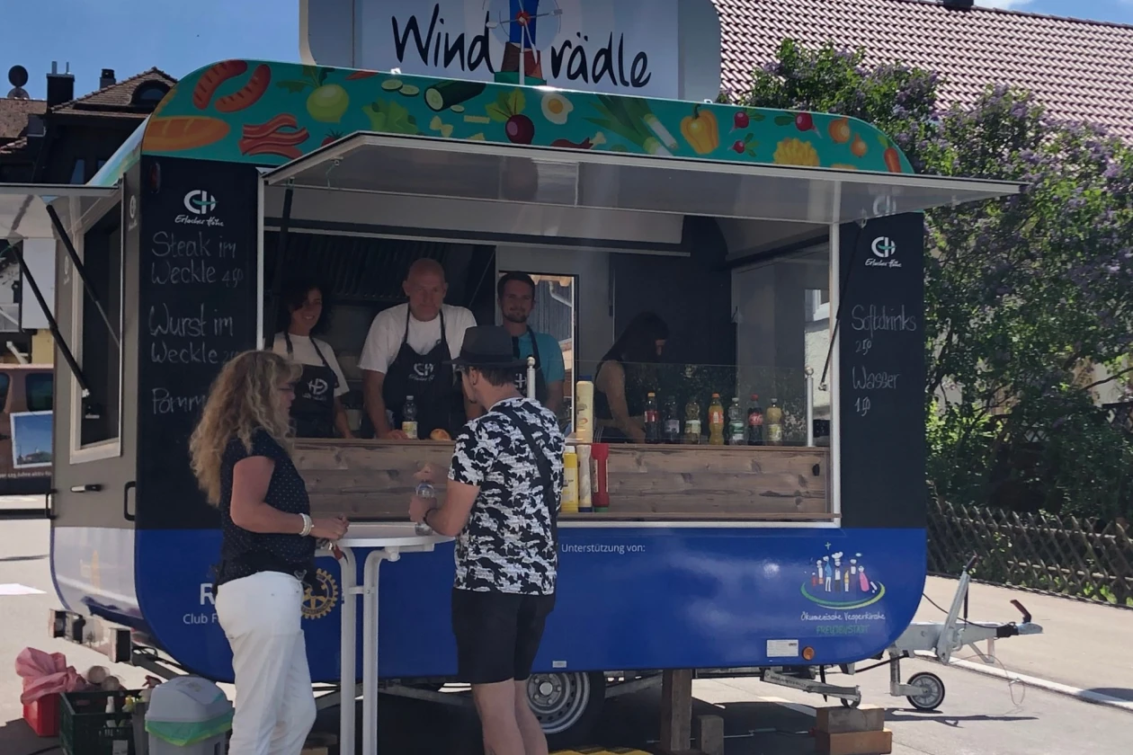 Zu sehen ist der Foodtruck "Windr&auml;dle" der Erlacher H&ouml;he, an dem zwei Personen Getr&auml;nke kaufen. Der Anh&auml;nger steht auf einem Parkplatz und zwei Personen mit Sch&uuml;rze stehen hinter dem Tresen.