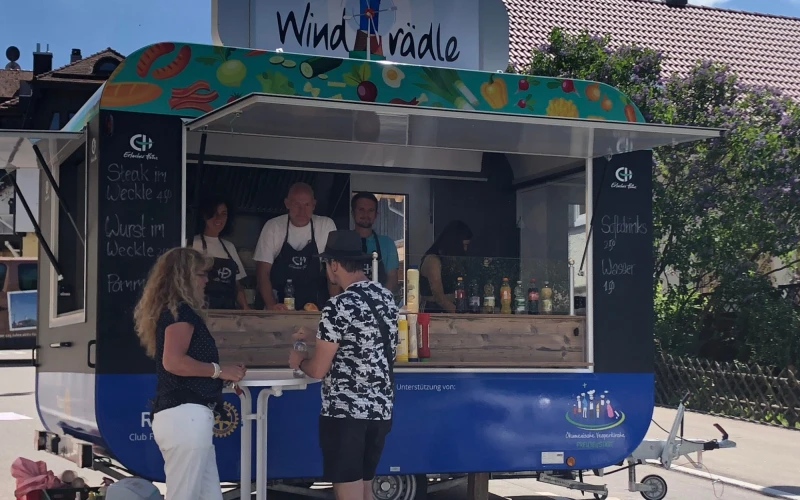 Zu sehen ist der Foodtruck "Windr&auml;dle" der Erlacher H&ouml;he, an dem zwei Personen Getr&auml;nke kaufen. Der Anh&auml;nger steht auf einem Parkplatz und zwei Personen mit Sch&uuml;rze stehen hinter dem Tresen.