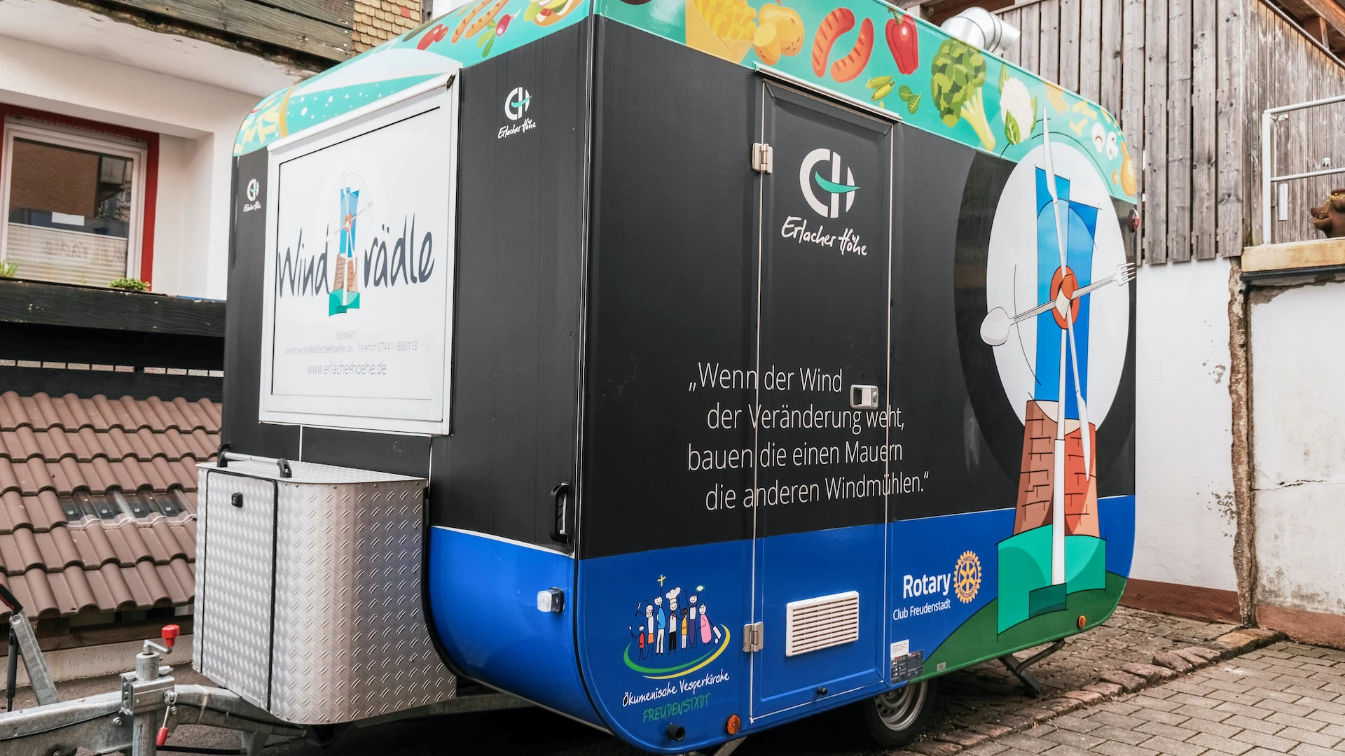 Ein mobiler Anh&auml;nger in Schwarz und Blau mit buntem Dach, beklebt mit der Aufschrift "Windr&auml;dle",  steht auf einem gepflasterten Hof.