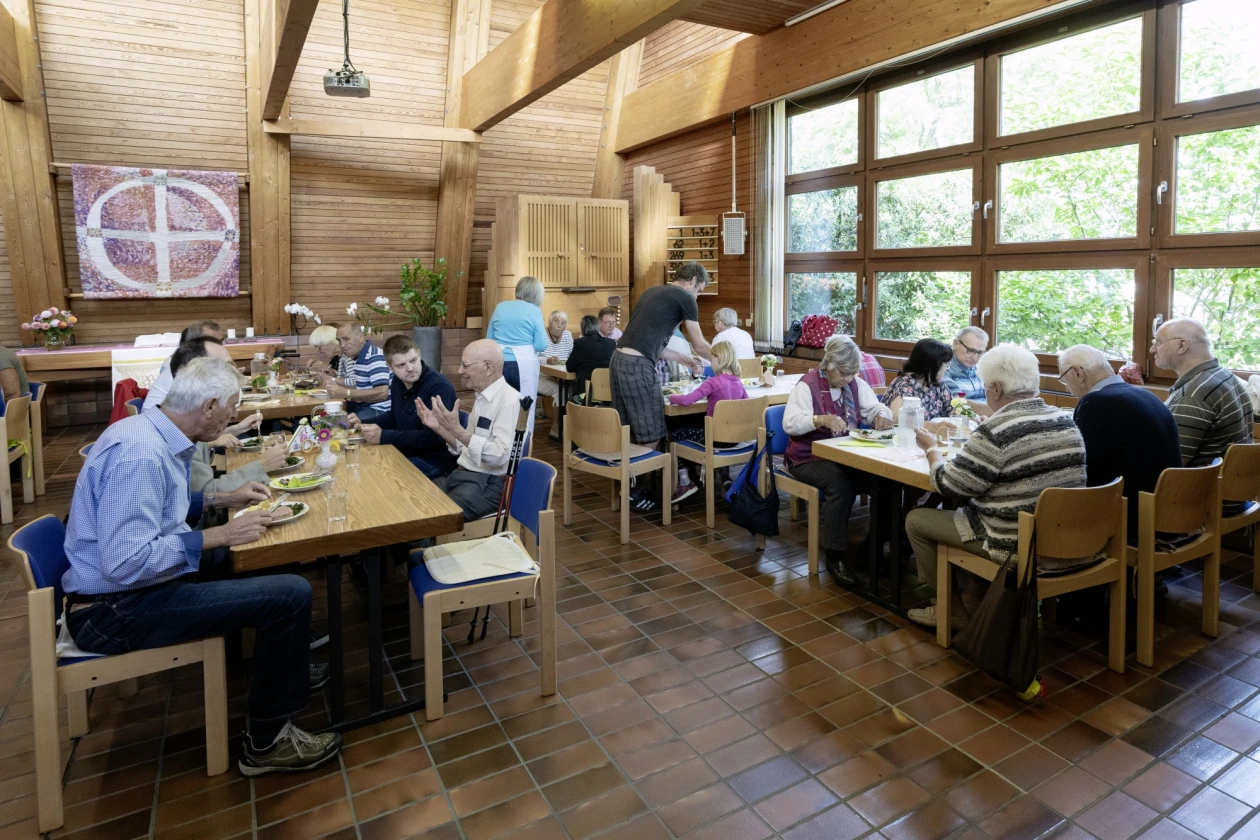 Menschen sitzen an Holztischen in einem hellen, holzverkleideten Raum mit gro&szlig;en Fenstern und essen gemeinsam Mittag. Der Raum ist freundlich und ger&auml;umig, mit Blick ins Gr&uuml;ne.