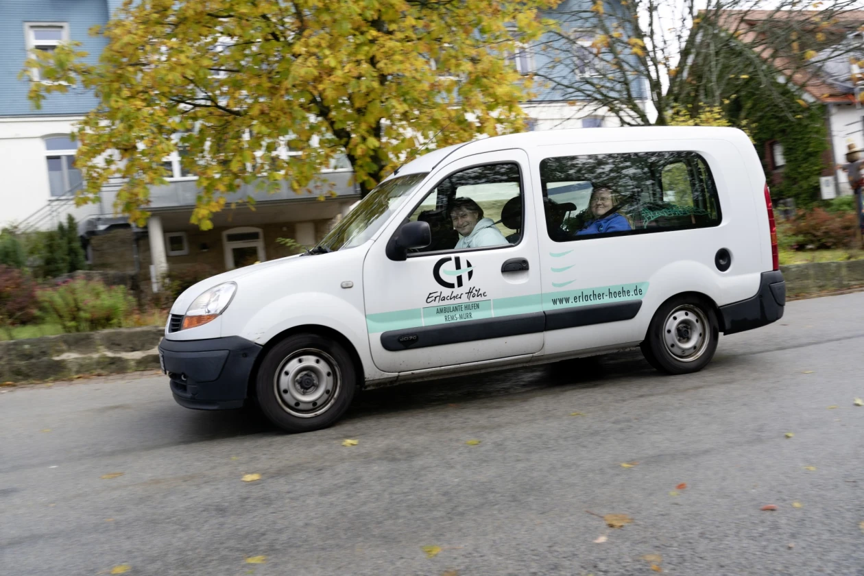 Wei&szlig;er Kastenwagen der Erlacher H&ouml;he auf einer Stra&szlig;e mit herbstlichem Baum im Hintergrund. Das Fahrzeug hat das Logo und die Beschriftung der Erlacher H&ouml;he seitlich angebracht. Zwei Personen sind im Auto zu sehen.
