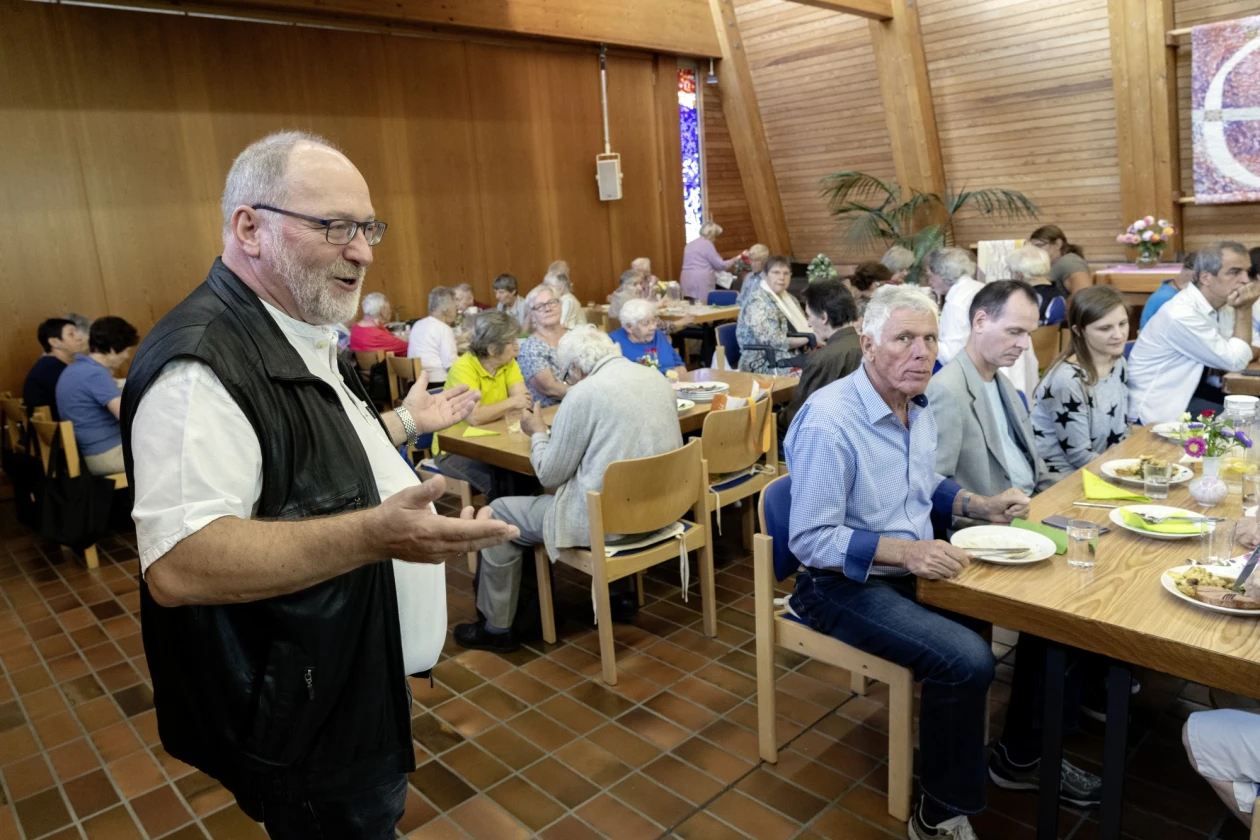Ein Mann mi einem wei&szlig;en T-Shirt, schwarzer Weste und Brille steht in einem Speisesaal mit vielen Menschen, die an Holztischen sitzen und essen. Der Raum hat Holzvert&auml;felungen und einen Steinboden.