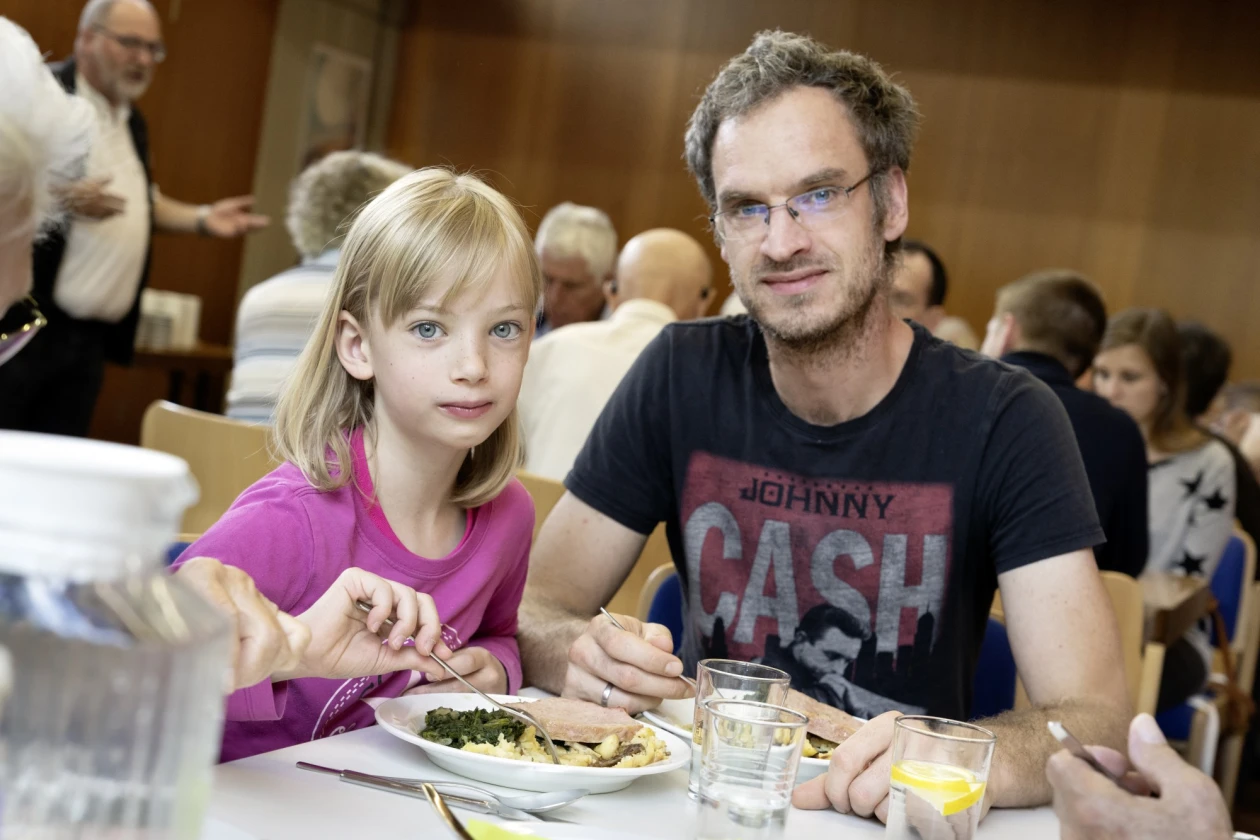 Ein Mann mit Johnny Cash T-Shirt und eine junges blondes M&auml;dchen sitzen gemeinsam an einem Tisch und essen. Sie haben Teller mit Essen vor sich und Wassergl&auml;ser auf dem Tisch, im Hintergrund sind weitere Personen zu sehen.