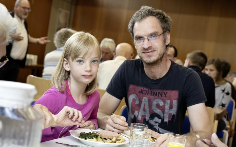 Ein Mann mit Johnny Cash T-Shirt und eine junges blondes M&auml;dchen sitzen gemeinsam an einem Tisch und essen. Sie haben Teller mit Essen vor sich und Wassergl&auml;ser auf dem Tisch, im Hintergrund sind weitere Personen zu sehen.