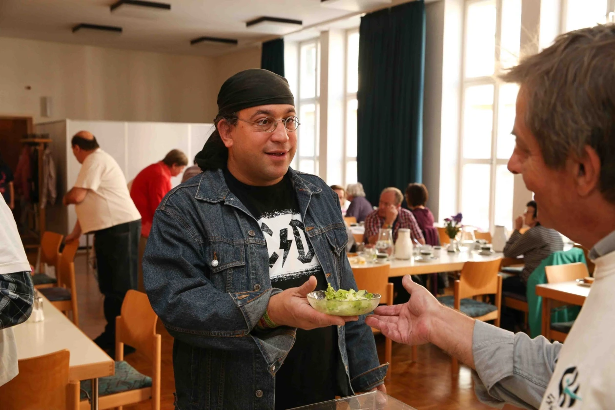 Ein Mann in Jeansjacke und schwarzer M&uuml;tze nimmt von einem anderen Mann in einem Speisesaal einen Teller mit Salat entgegen, w&auml;hrend im Hintergrund weitere Personen am Essen sind.