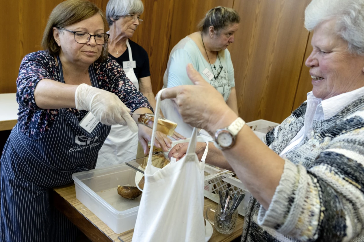 Zwei &auml;ltere Frauen in Sch&uuml;rzen arbeiten gemeinsam an einer Essensausgabe, eine tr&auml;gt Plastikhandschuhe und legt mit einer Zange Geb&auml;ckst&uuml;cke in die Stofftasche einer &auml;lteren Dame.
