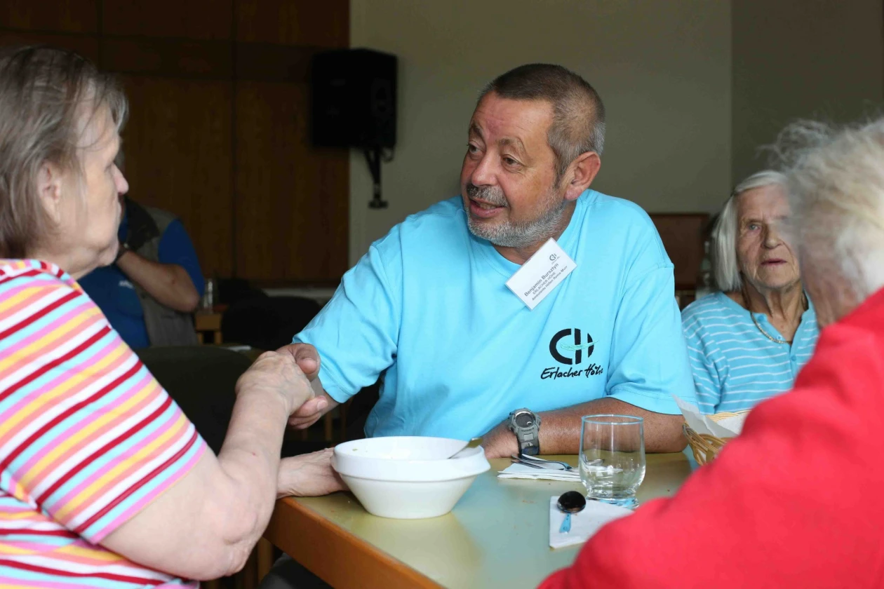 Mehrere Personen sitzen an einem Tisch mit Sch&uuml;sseln und Getr&auml;nken, ein Mann in einem Shirt mit dem Erlacher H&ouml;he Logo sch&uuml;ttelt die Hand eine Frau.