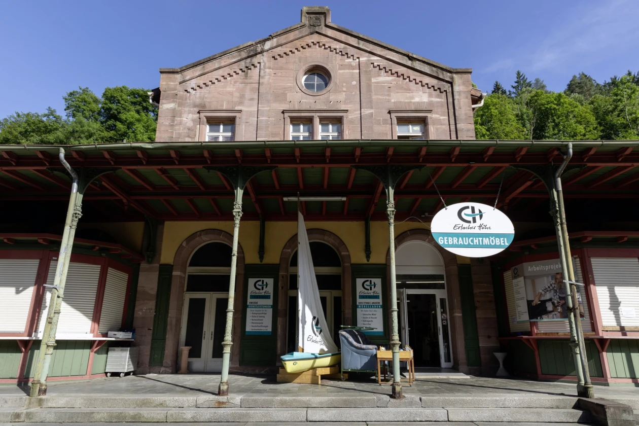 Ein Geb&auml;ude der Erlacher H&ouml;he mit Backsteinfassade, &uuml;berdachter Veranda und Eingangstreppe, gelegen vor einem gr&uuml;nen Waldhintergrund. Vor dem Eingang stehen ein Segelboot sowie ein Schild mit der Aufschrift "Gebrauchtm&ouml;bel" und dem Logo der Erlacher H&ouml;he.