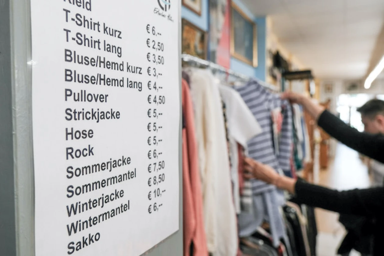 Preisliste f&uuml;r Kleidungsst&uuml;cke in einem Kleidungsgesch&auml;ft mit verschiedenen Artikeln wie T-Shirts, Pullover und Jacken. Die Preise sind in Euro angegeben, und im Hintergrund sind Kleidungsst&uuml;cke und zwei Personen zu sehen, die das Angebot durchst&ouml;bern.