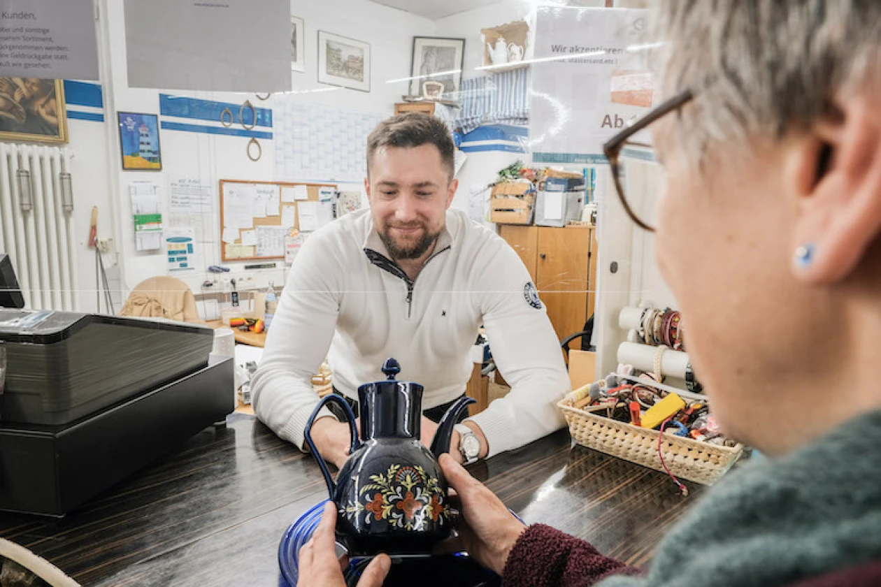 Ein Mann in einem wei&szlig;en Pullover steht l&auml;chelnd hinter der Kasse und blickt auf die Teekanne, die eine Frau vor ihm in den H&auml;nden h&auml;lt. Im Hintergrund sind Pinnw&auml;nde und Bilder an der Wand zu sehen.
