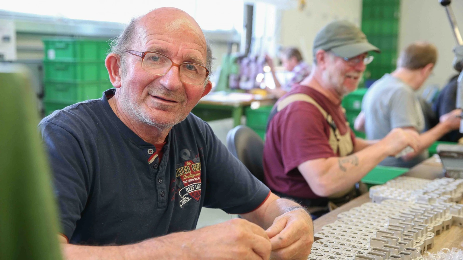 Ein &auml;lterer Mann mit Brille l&auml;chelt in die Kamera, w&auml;hrend er an einem Arbeitsplatz sitzt. Er tr&auml;gt ein schwarzes T-Shirt mit Aufdruck und arbeitet in einer Werkstatt mit weiteren Personen im Hintergrund.