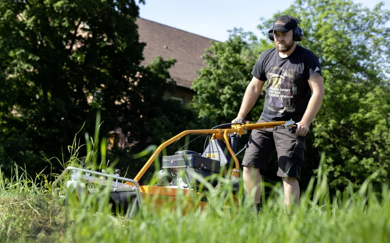 Ein Mann in Arbeitskleidung m&auml;ht Gras mit einem gelben Rasenm&auml;her an einem sonnigen Tag.