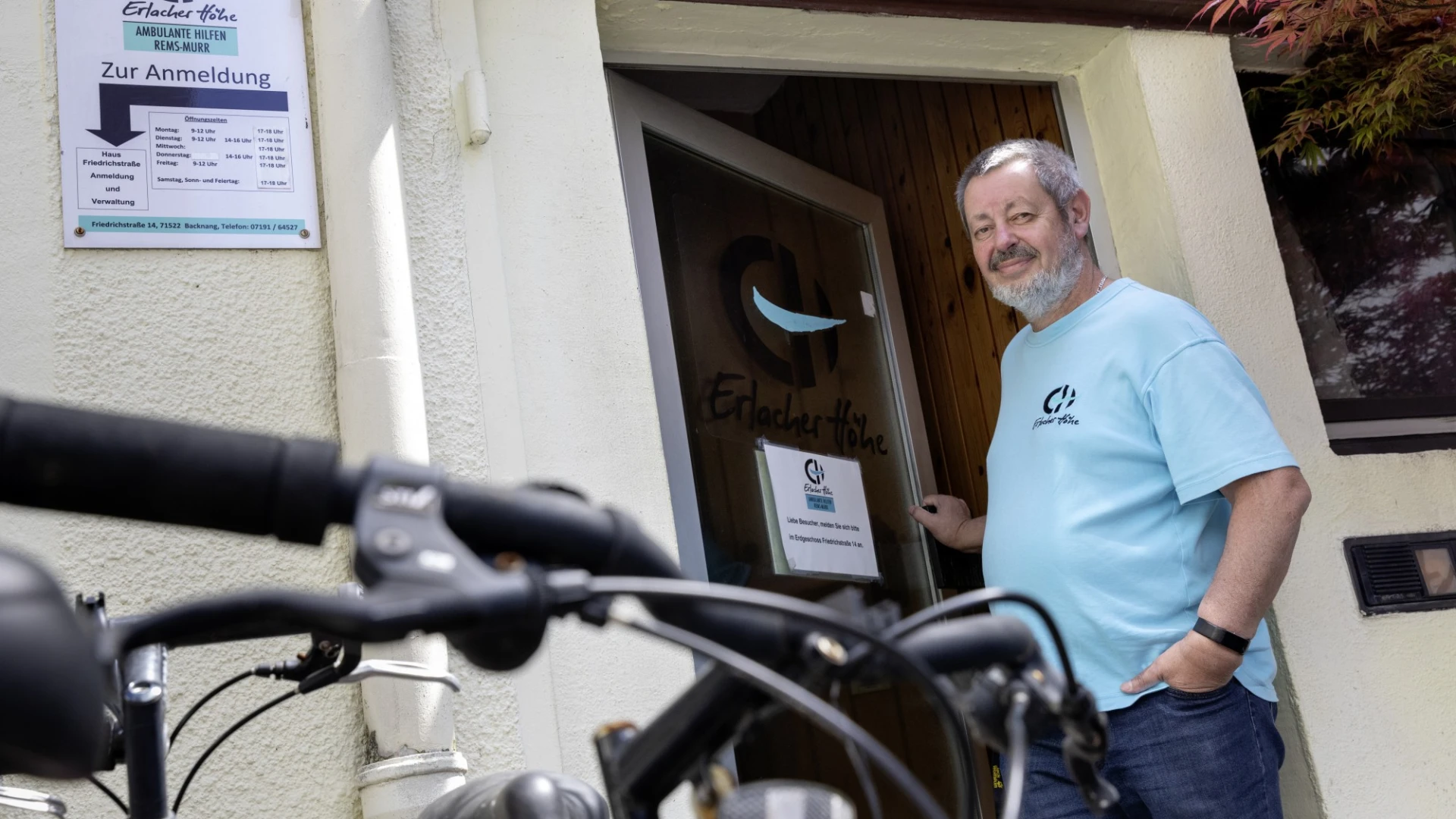 Ein Mann in einem T-Shirt mit Erlacher H&ouml;he Logo steht l&auml;chelnd an der Eingangst&uuml;r eines Geb&auml;udes, im Vordergrund ist ein Fahrradlenker zu sehen, daneben h&auml;ngt ein Schild von der Erlacher H&ouml;he mit dem Aufschrieb: "Ambulante Hilfen Rems-Murr".