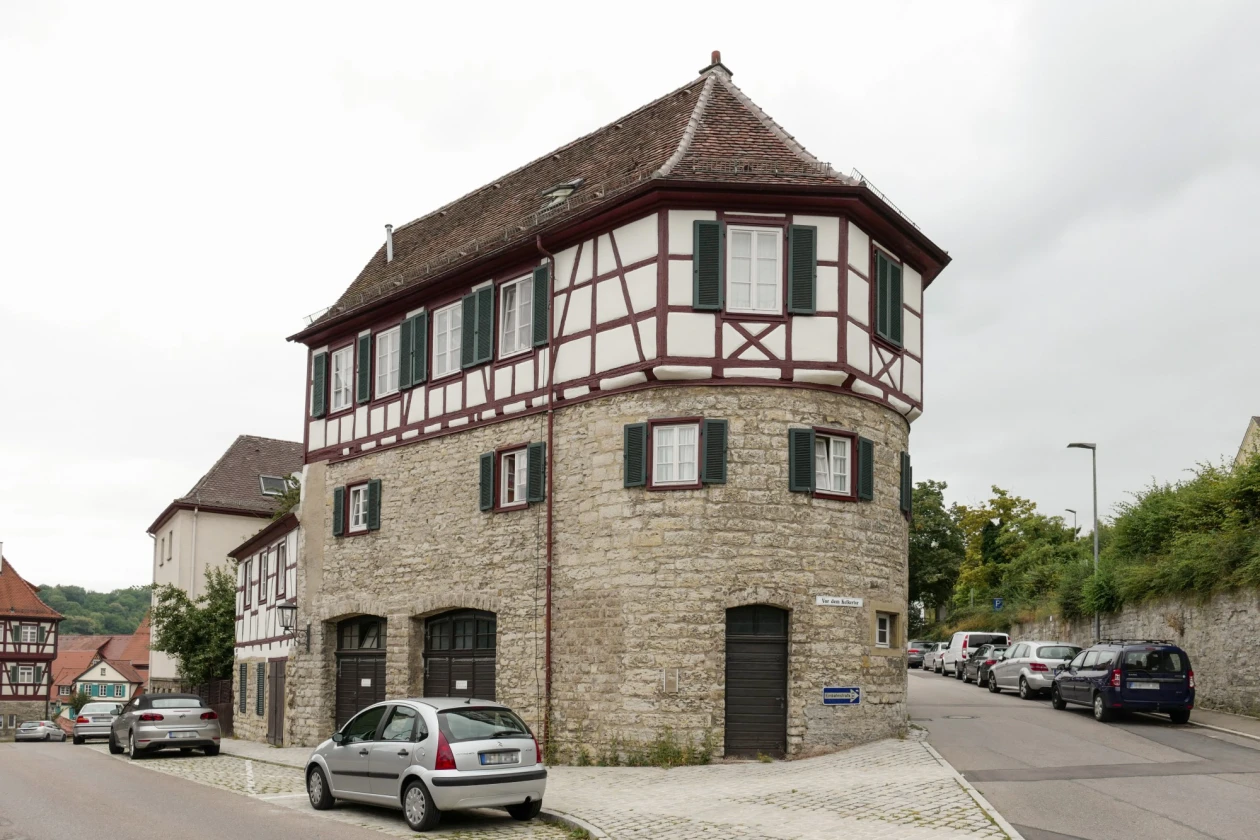 Ein Fachwerkhaus mit Natursteinunterbau an einer Stra&szlig;enecke, umgeben von parkenden Autos und weiteren Fachwerkh&auml;usern im Hintergrund. Das markante Geb&auml;ude hat einen turmartigen Grundriss.