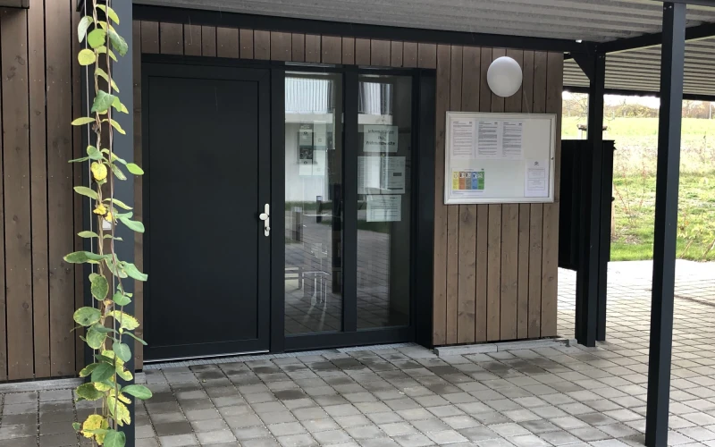 Moderner Eingangsbereich mit Holzfassade, schwarzer T&uuml;r mit Glasfenstern und einem &uuml;berdachten Vorbau. Links w&auml;chst eine Kletterpflanze mit gelben und gr&uuml;nen Bl&uuml;ten an der Wand. Ein Informationsschild h&auml;ngt rechts neben der Eingangst&uuml;r.