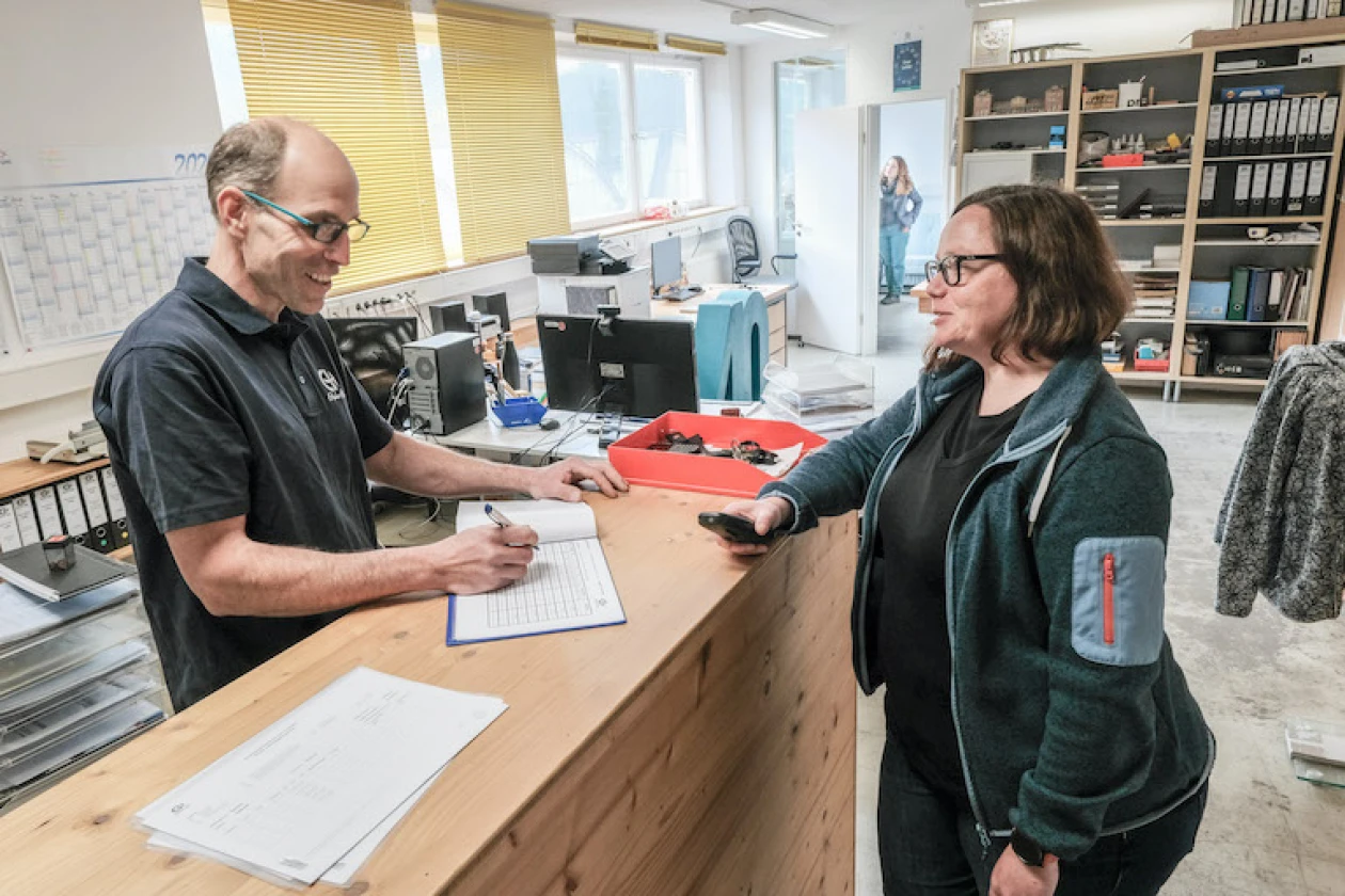 Ein Mitarbeiter steht hinter einem Holztresen und hat einen Kugelschreiber und ein Dokument in der Hand. Vor ihm steht eine Frau mit einem Telefon in der Hand. Beide tragen Brillen und arbeiten in einem hellen B&uuml;ro mit Regalen und Aktenordnern im Hintergrund.