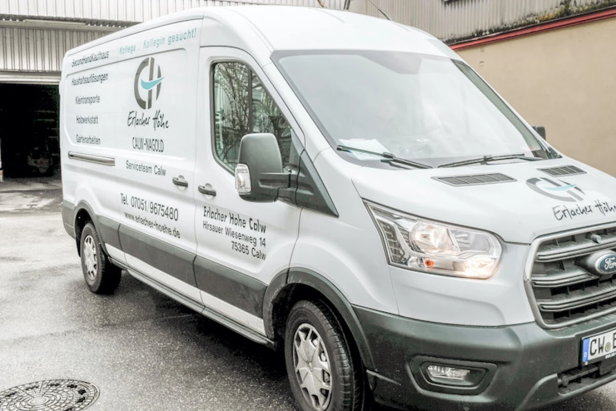 Grauer Transporter mit Firmenlogo und Kontaktinformationen von der Erlacher H&ouml;he, geparkt auf einem Hinterhof mit Betonboden und Geb&auml;uden im Hintergrund.
