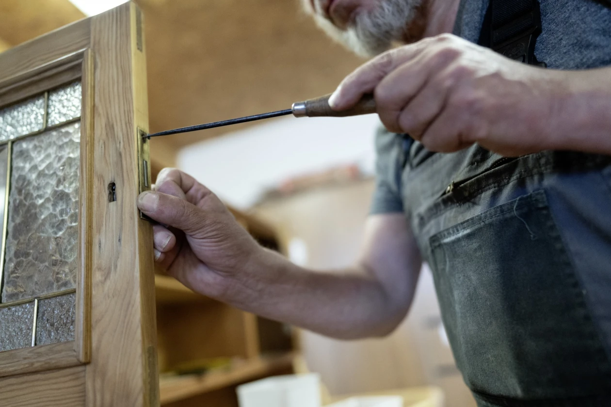 H&auml;nde bei Reparaturarbeit an einem Holzrahmen, eine Hand h&auml;lt einen Schraubendreher und befestigt ein Scharnier an der Holzoberfl&auml;che.